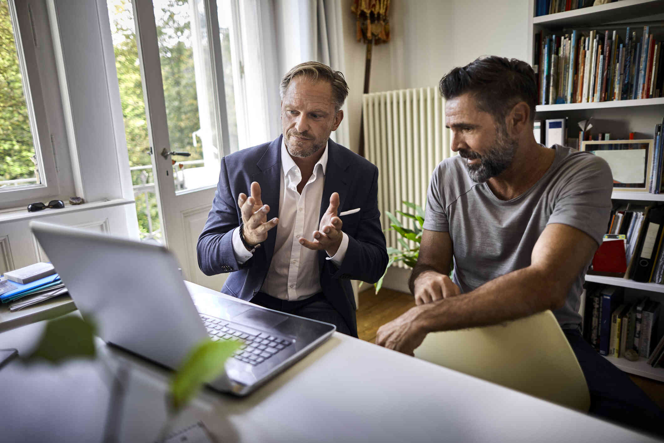 A man in a suit gestures towards a laptop sitting on a desk in front of him. Another man in casual attire sits next to him with a focused expression.